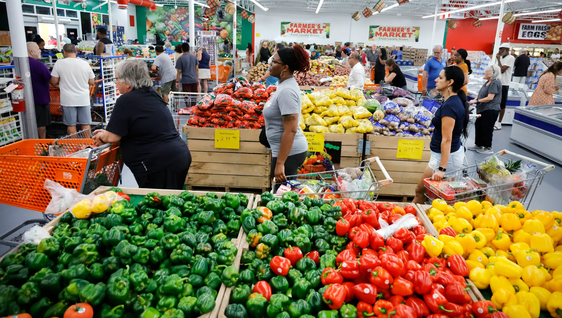 Shoppers browse bountiful displays of fresh produce, including red and green bell peppers, yellow squash, and other fruits and vegetables, at an indoor 99 Cents Farmers Market.